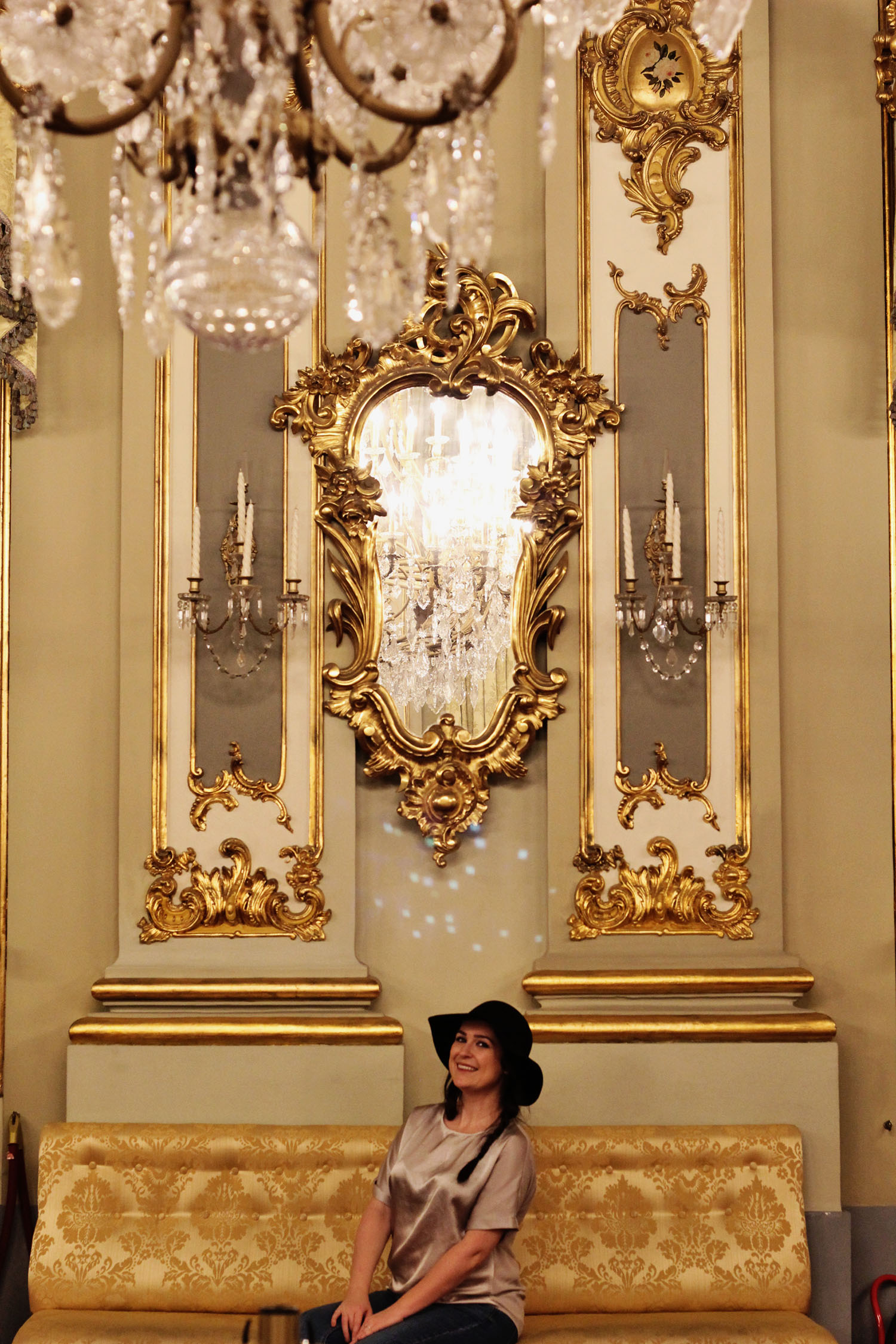 woman wears black floppy hat sitting on an ornate sofa in an ornate gold room