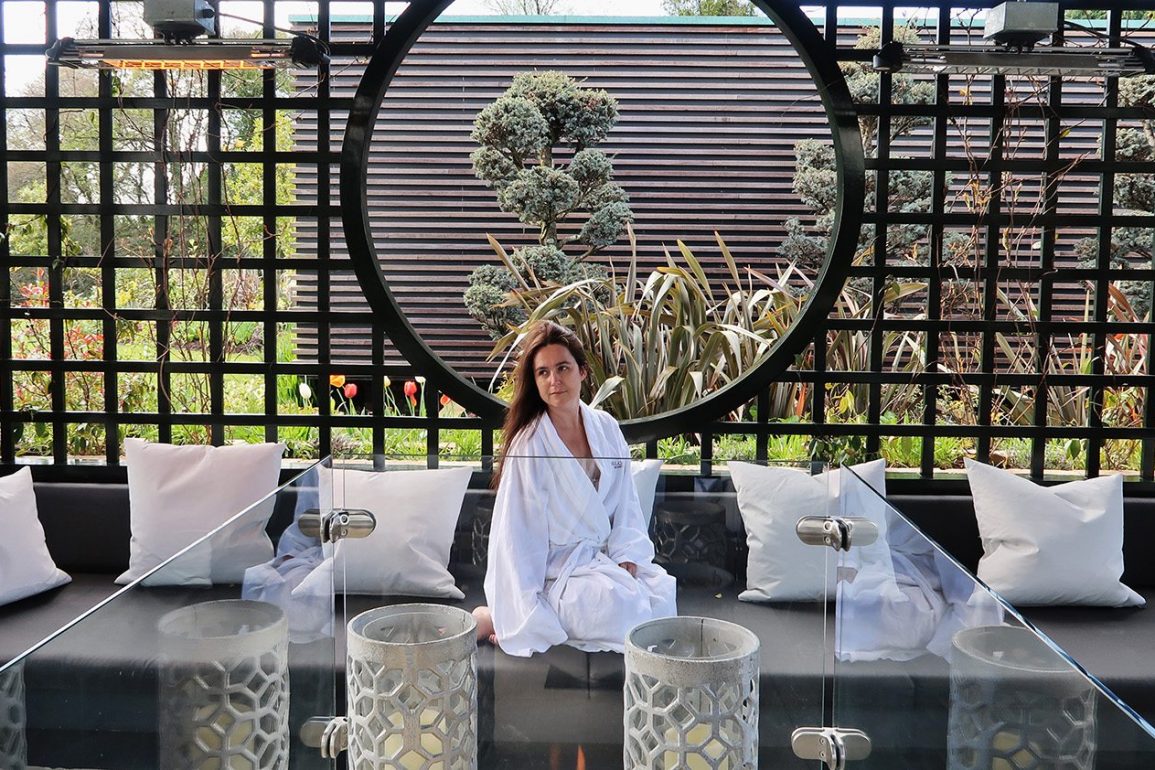 Woman in a white spa robe sits in an outside seating area with a modern firepit in the foreground
