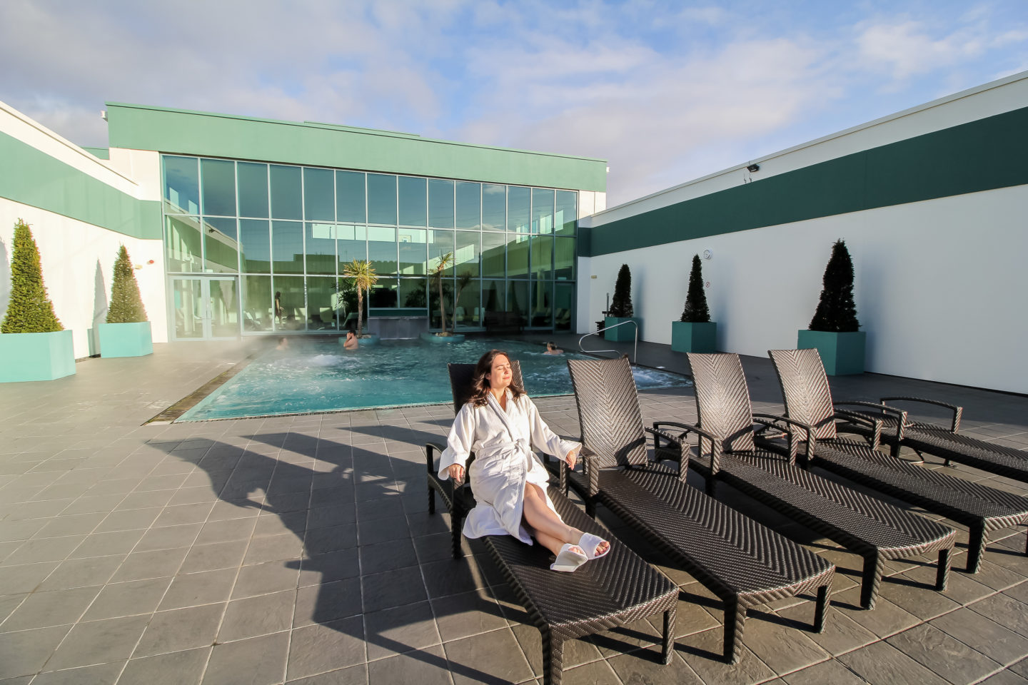 woman in white dressing gown lies on sun bed in front of a pool at the spa