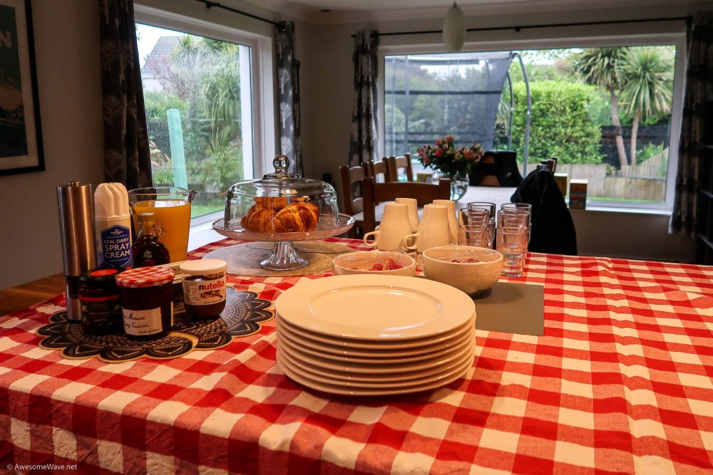 kitchen top with red and white checked table cloth and piles of plates, mugs and glasses. There are croissants and a selection of jams and creams.