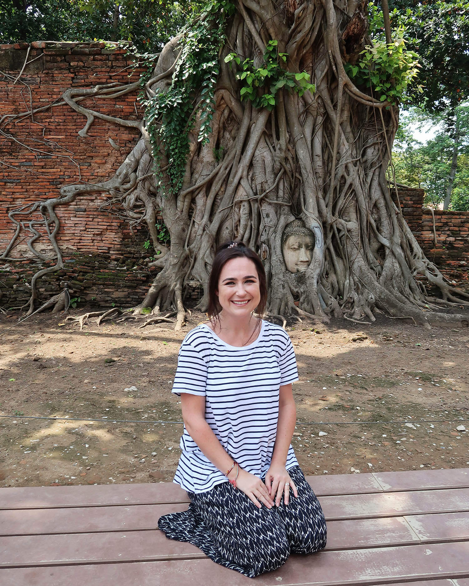 Photo of woman kneeling on wooden platform in front of a tree with large visible roots and the statue of a buddha head
