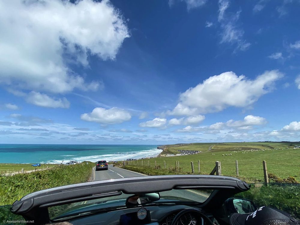 blue skies with white fluffy clouds, a winding road with a car in front and the open top of the car just visibly.